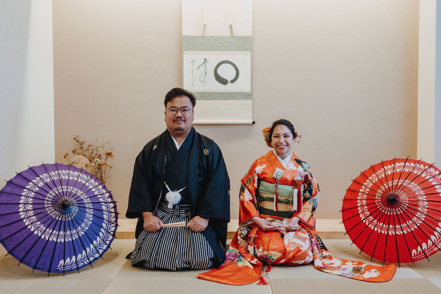Couple in traditional Japanese wedding attire seated with decorative umbrellas indoors.