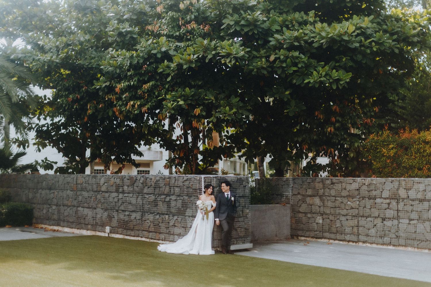 A wedding couple stands in front of a stone wall and trees in an elegant outdoor garden setting.