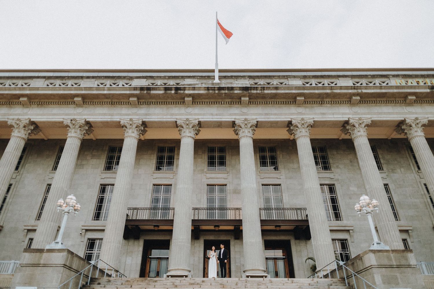 Grand neoclassical building with white columns and a red flag atop, with wedding couple on stairs.