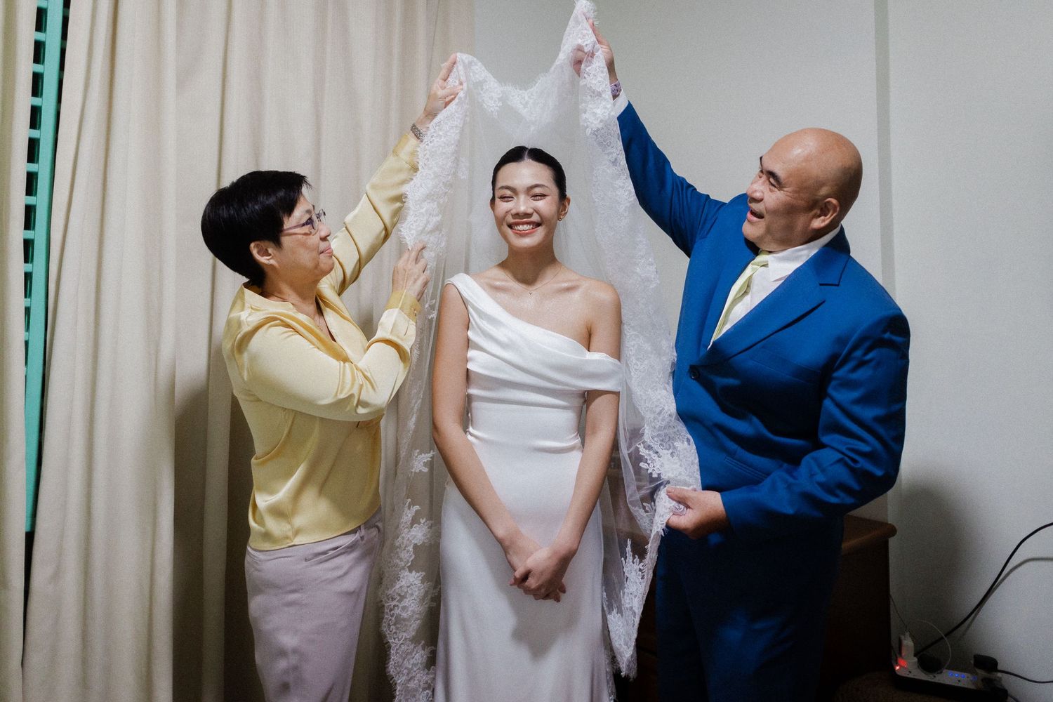 Bride in white wedding gown smiles as two family members help lift her veil indoors.
