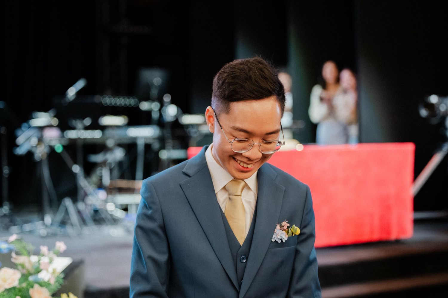 Smiling groom in blue suit with gold tie and boutonniere standing on stage at wedding ceremony.