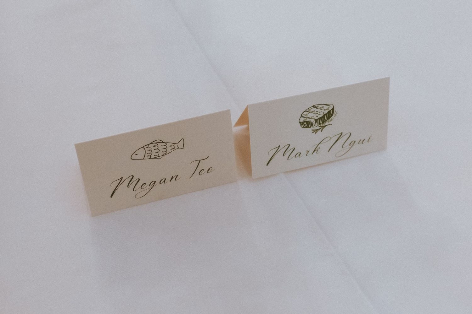 Elegant wedding place cards for Megan Tee and Mark Ngau with gold script and food icons on white background.