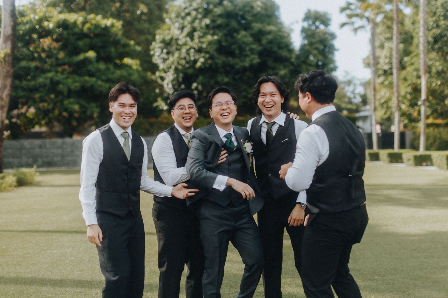 Groomsmen in black vests and ties share a candid laughing moment together on a sunny lawn.