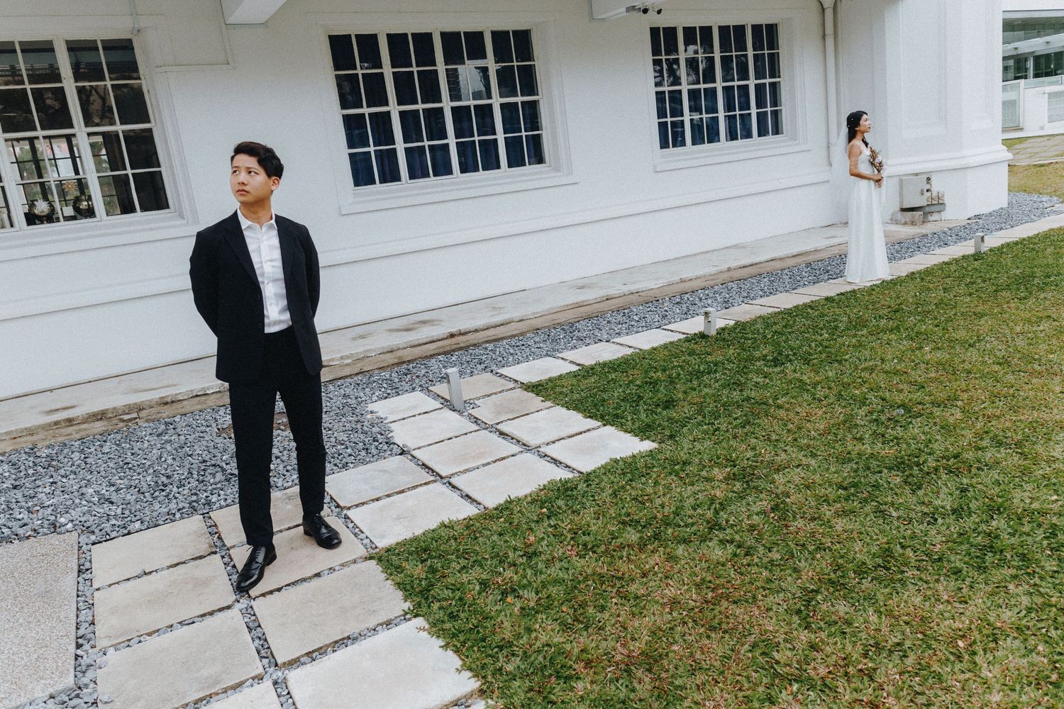 Two people in contrasting outfits pose separately outside a white building with windows.