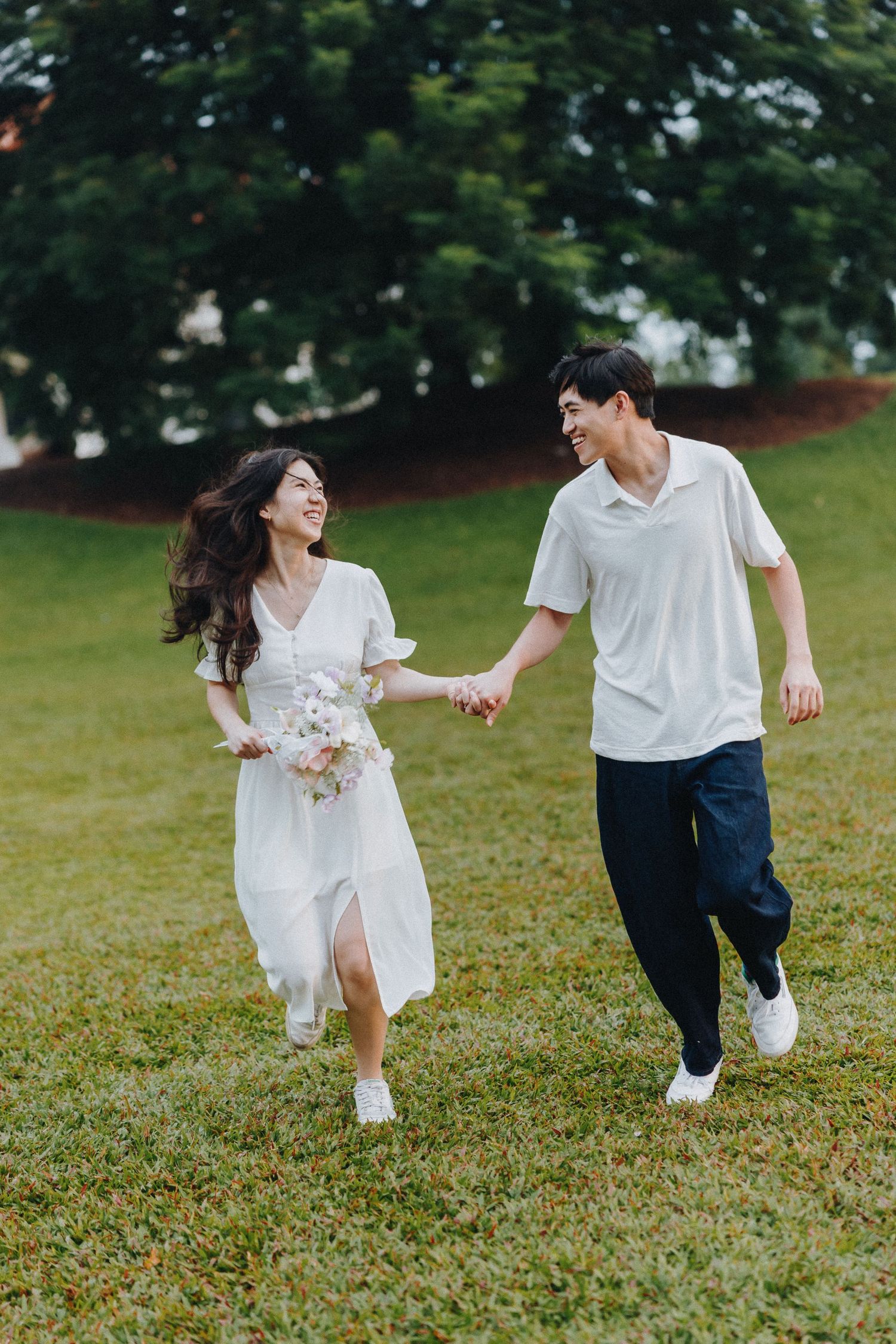 A couple in white outfits runs playfully through a green field on a sunny day.