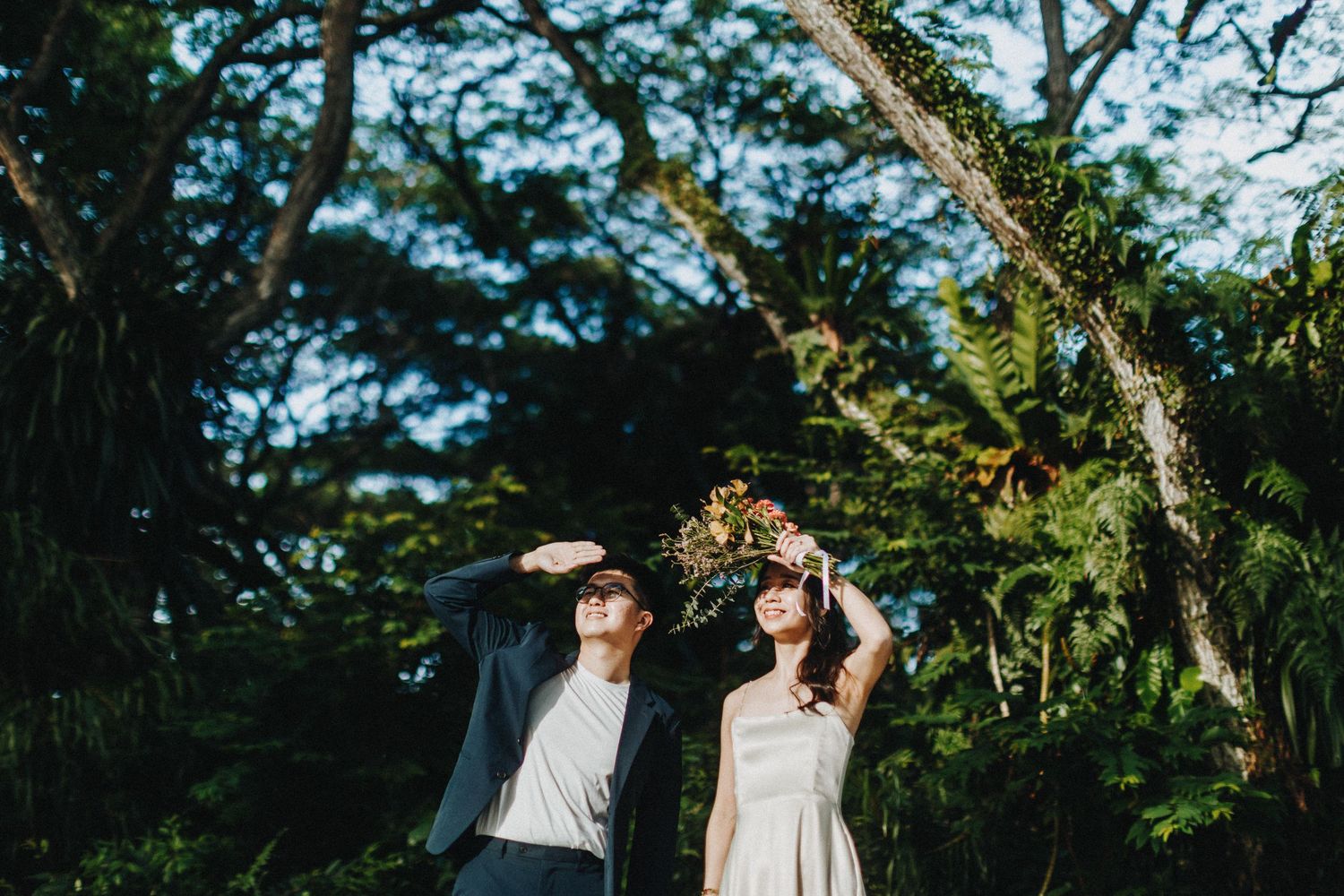Wedding couple poses playfully under lush green trees in an outdoor garden setting in natural lighting.