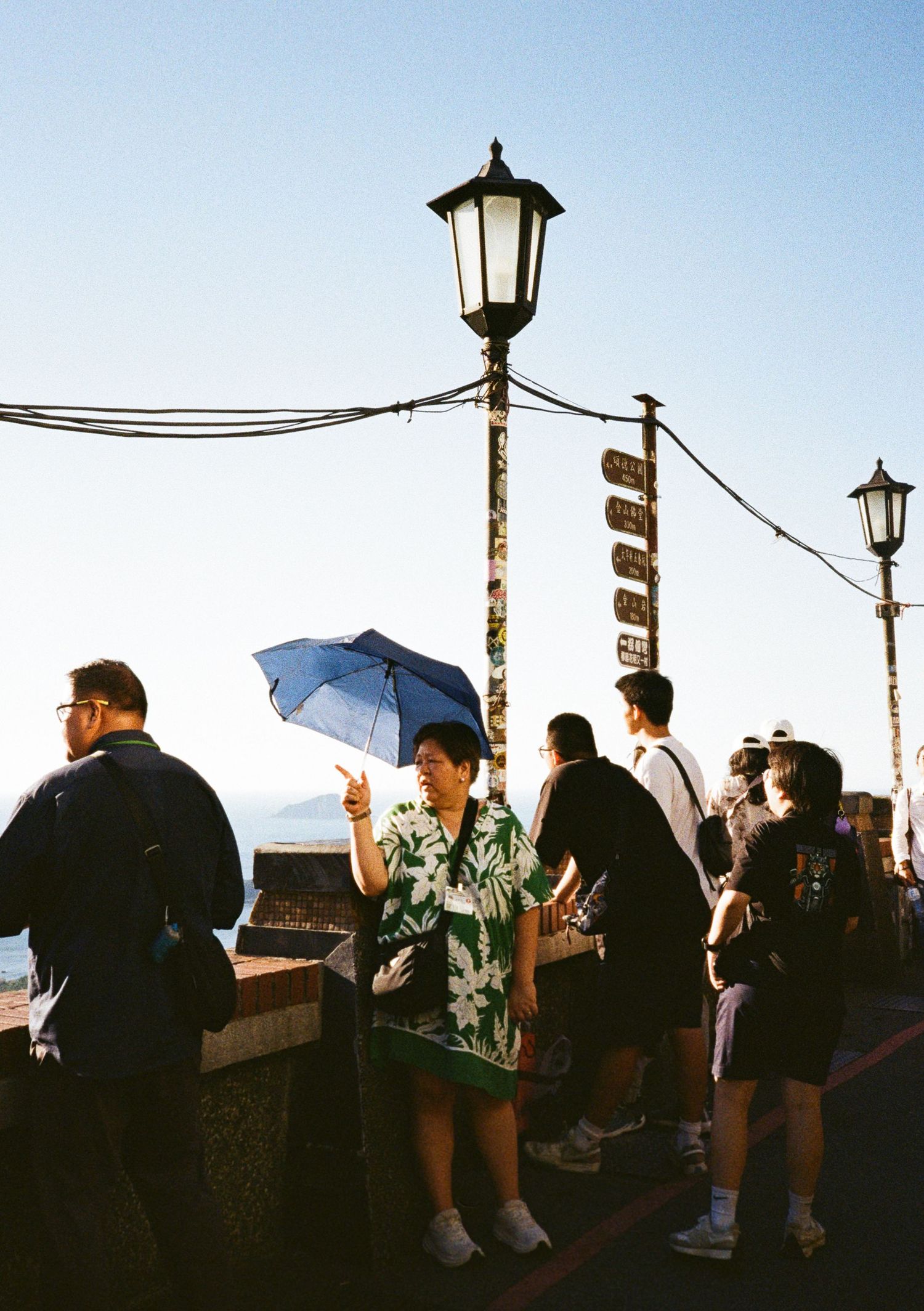 People gather along a seaside boardwalk at dusk, silhouetted against the sky with vintage lamp posts.