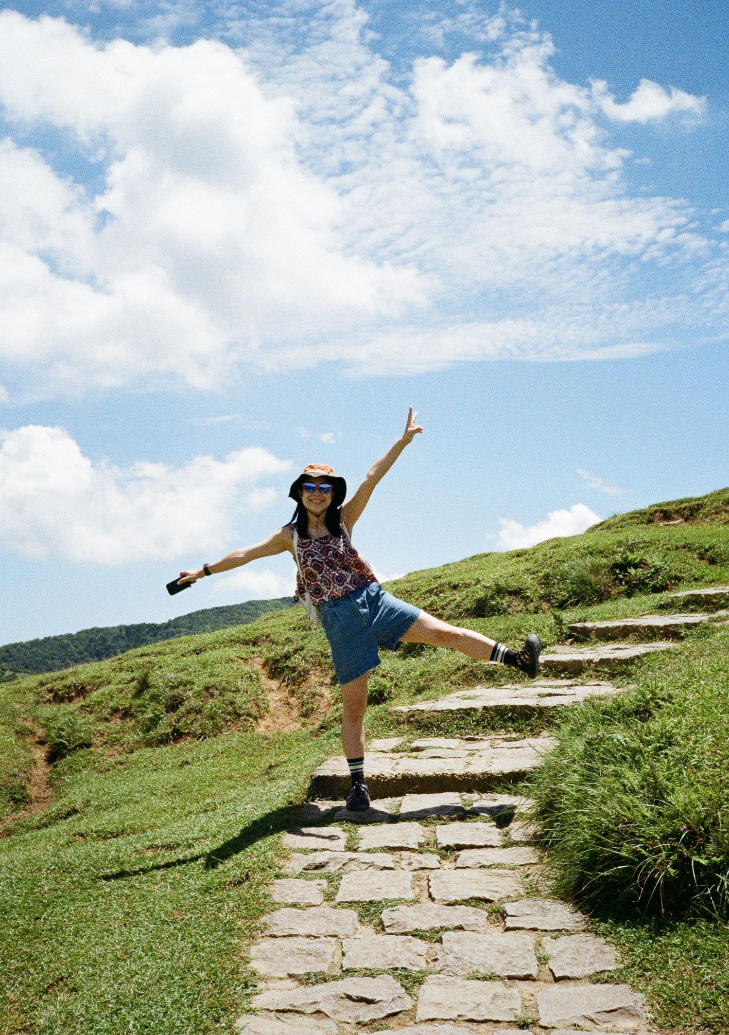 A series of joyful jumps and poses on a stone hiking trail winding through green mountain hills on a sunny day.
