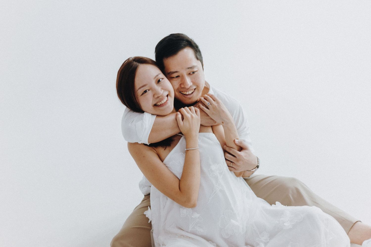Couple in white outfits embrace lovingly against a light background during a romantic photoshoot.