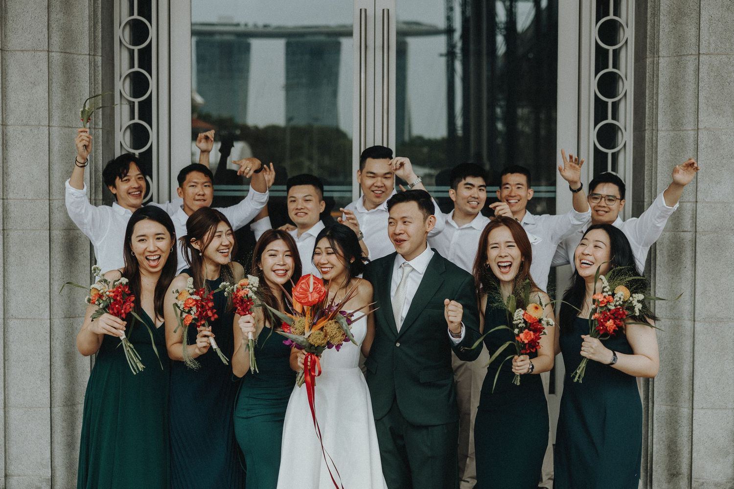 Wedding party celebrates joyfully on the steps of a grand columned building with colorful bouquets.