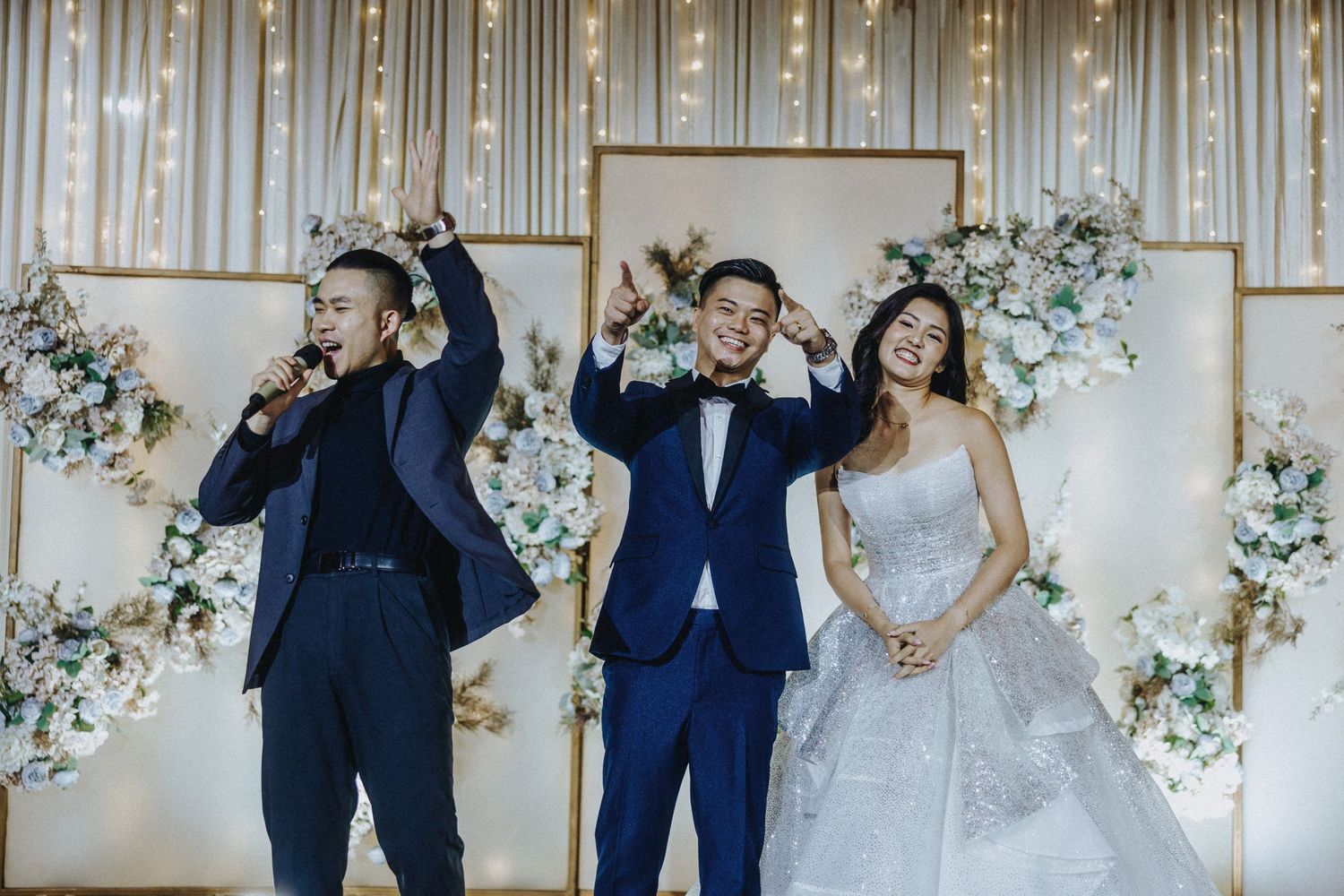Wedding celebration scene with people raising hands against a decorated backdrop with string lights and flowers.