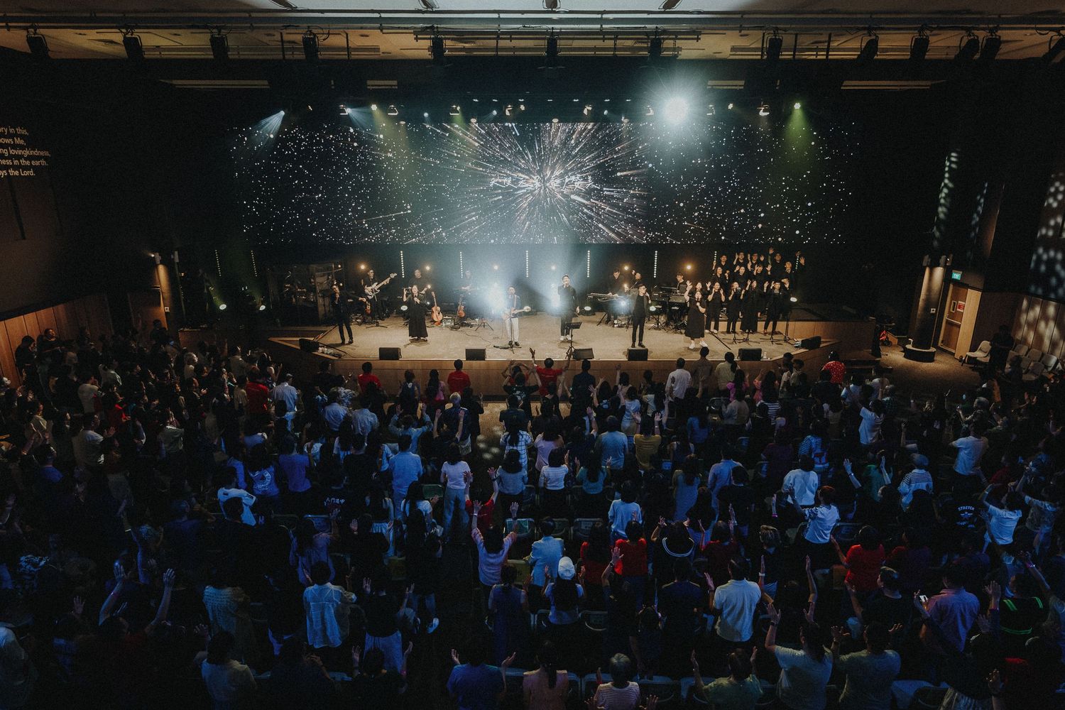 Concert venue with large crowd and spotlights illuminating a stage during a live performance.