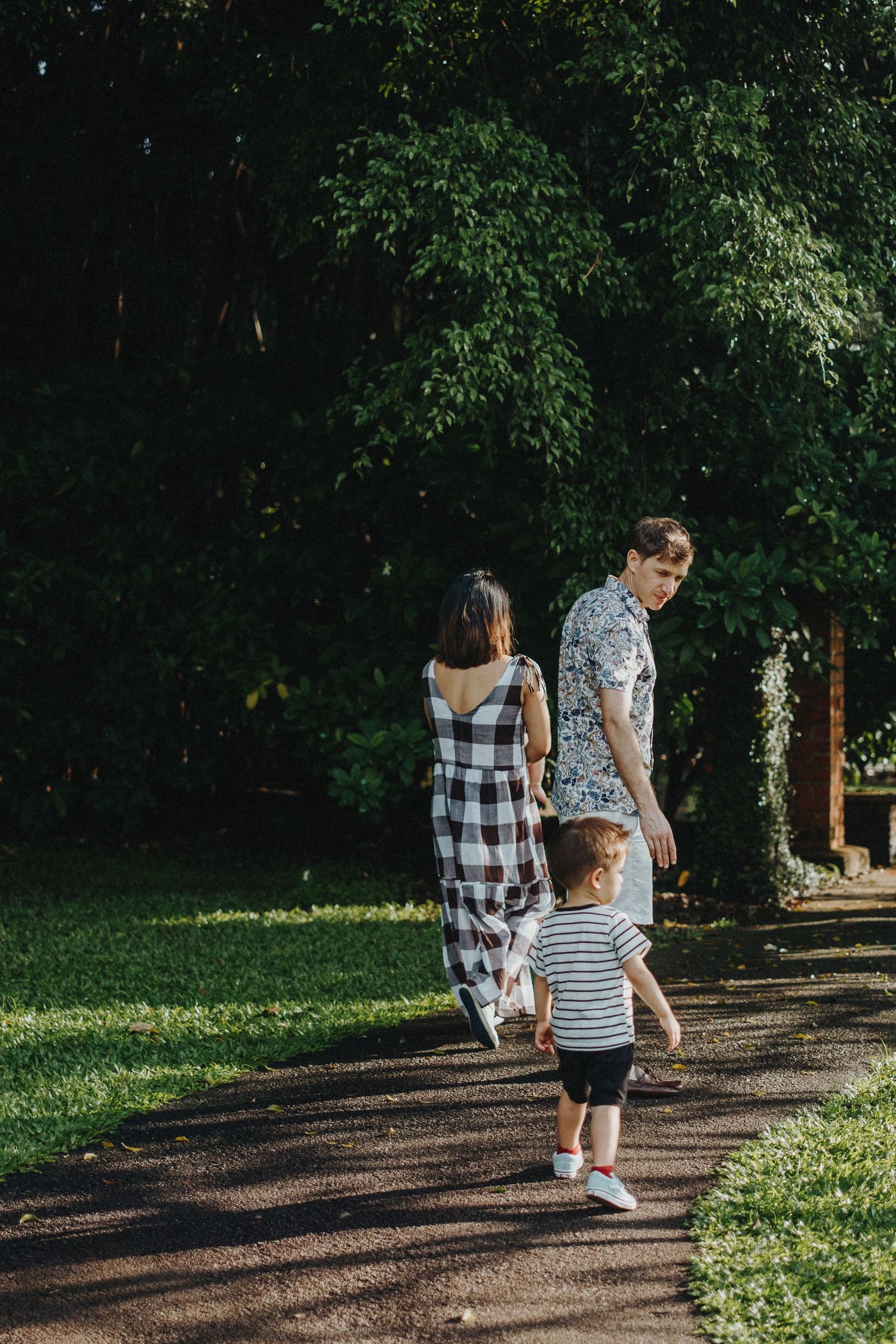 Family walks along tree-lined path in park during sunny afternoon wearing casual summer clothes.