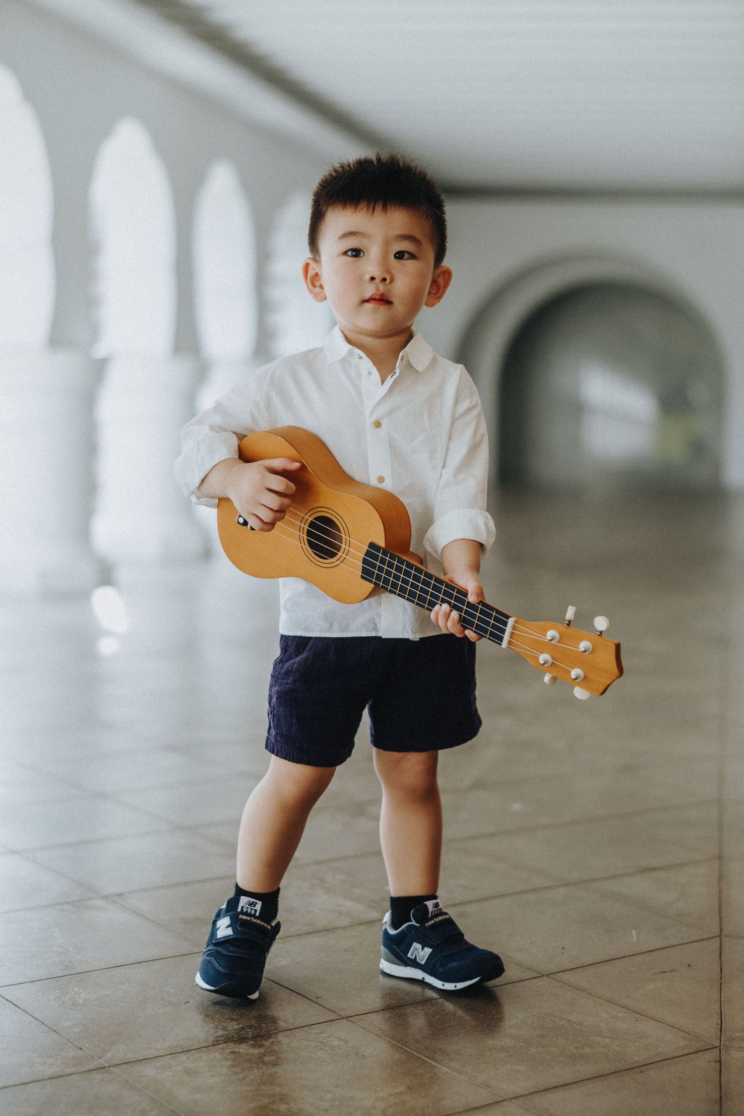 A young child wearing a white shirt and dark shorts holds a ukulele while standing in an arched hallway.
