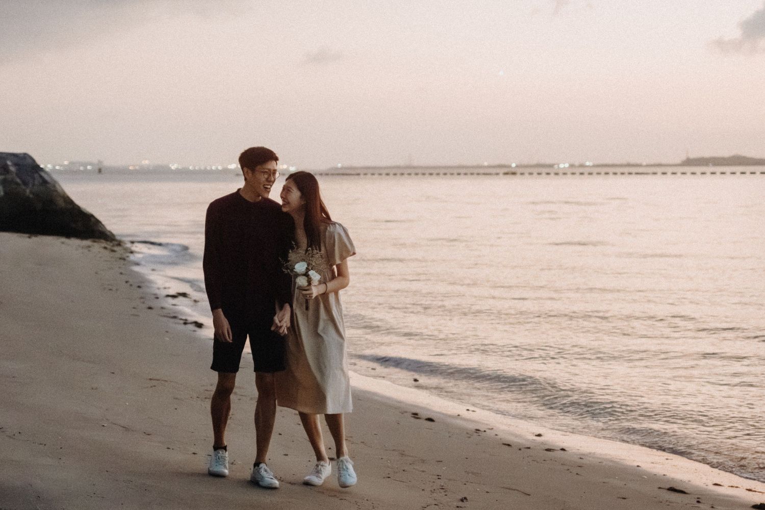 Two people walk together along a peaceful beach shoreline at sunset while ocean waves gently roll in.