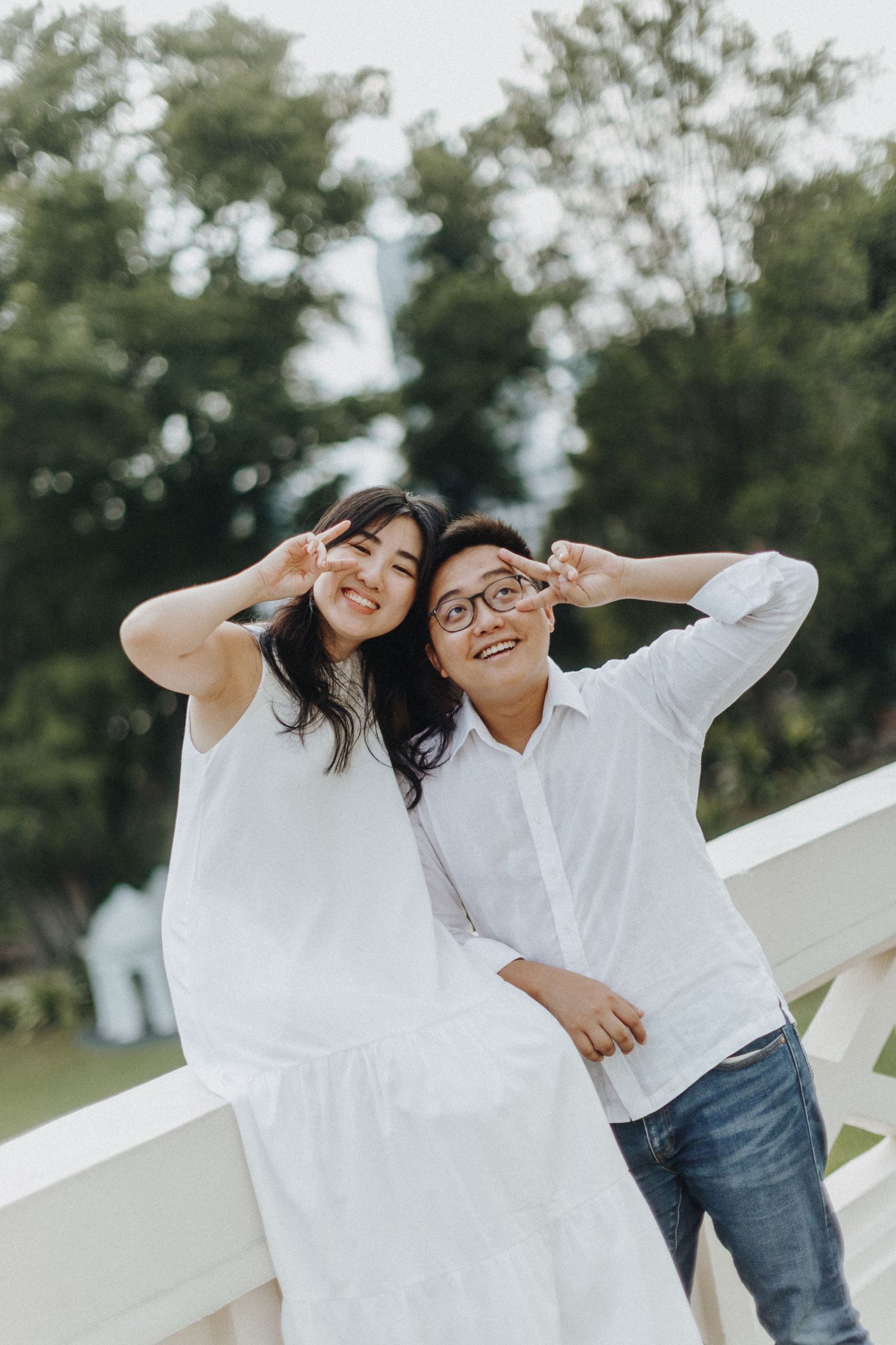 A joyful couple in matching white outfits make peace signs while posing outdoors against a natural background.
