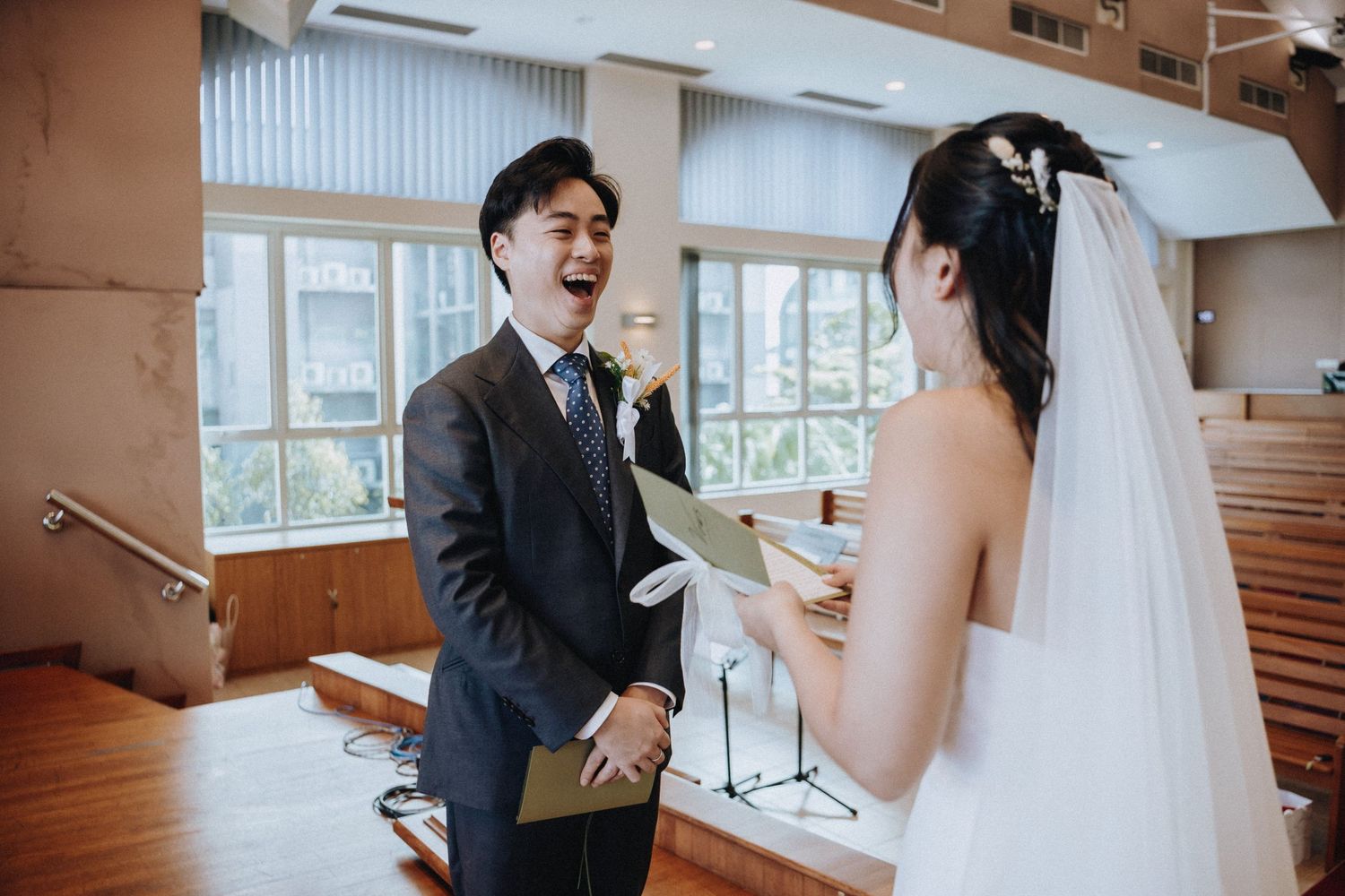Groom laughs joyfully as bride reads wedding vows during church ceremony.