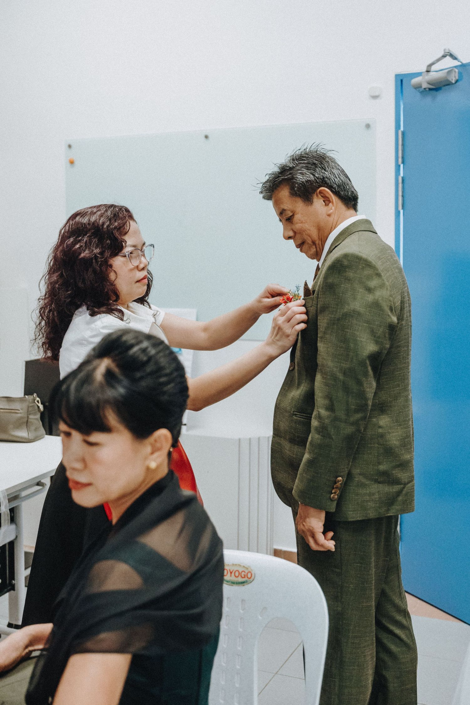 A woman adjusts a boutonniere on a man in a green suit while another woman sits in the foreground.