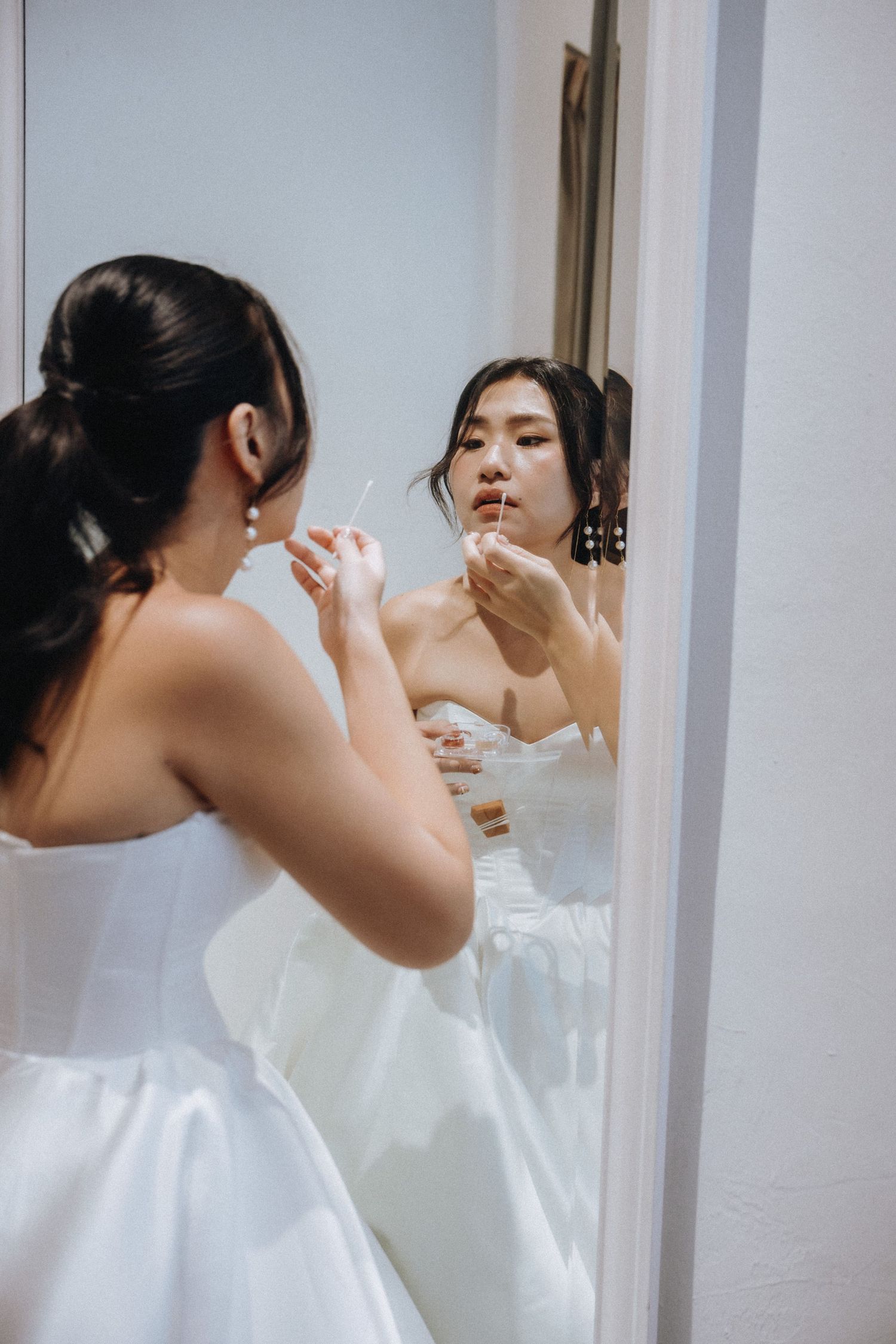 Asian bride in strapless white gown applies lip gloss while looking in mirror during wedding preparation.