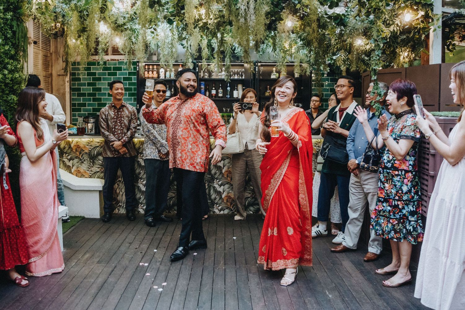 Couple in red Indian wedding attire celebrate with guests on decorated outdoor deck.