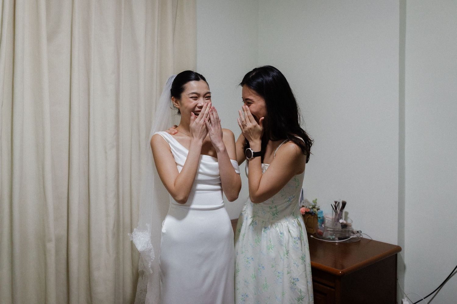Bride in white gown and veil laughing with bridesmaid in floral dress, both covering mouths in surprise.