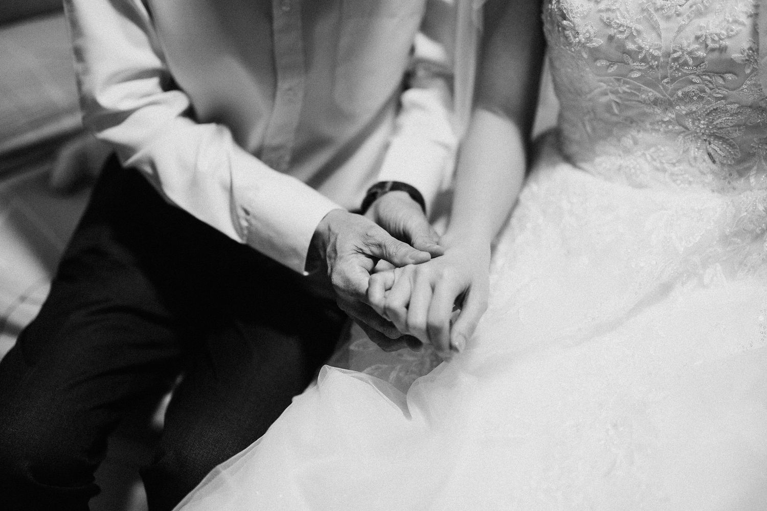 A black and white close-up of hands clasped together, with a wedding dress visible in the frame.