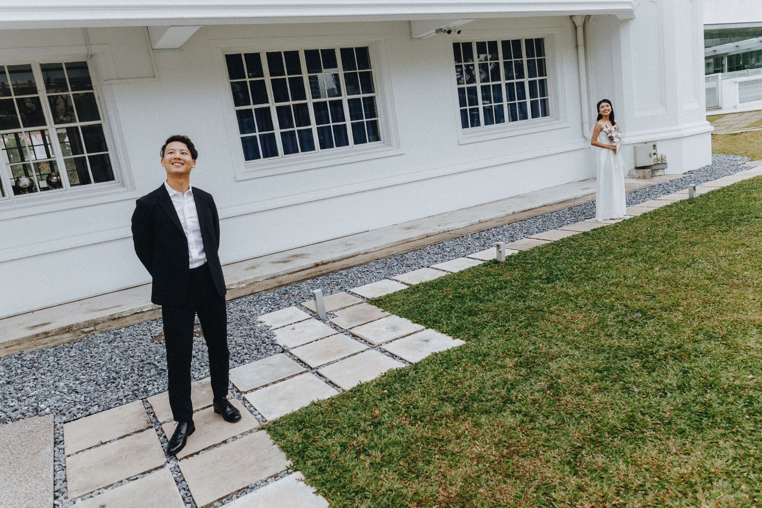 A couple poses separately outside a white building with windows and a stone path.