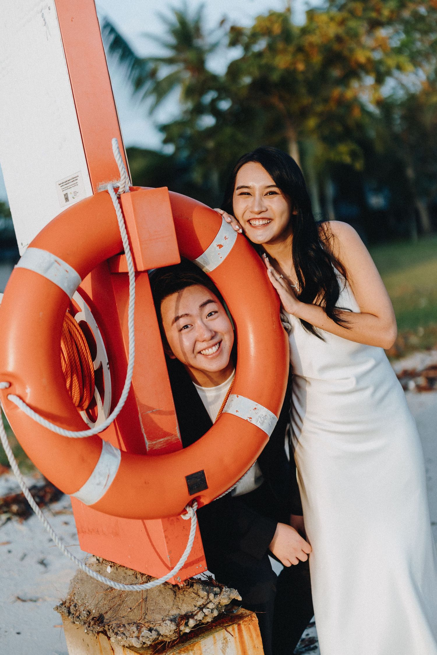A couple playfully poses with a bright orange life ring on a beach with palm trees.