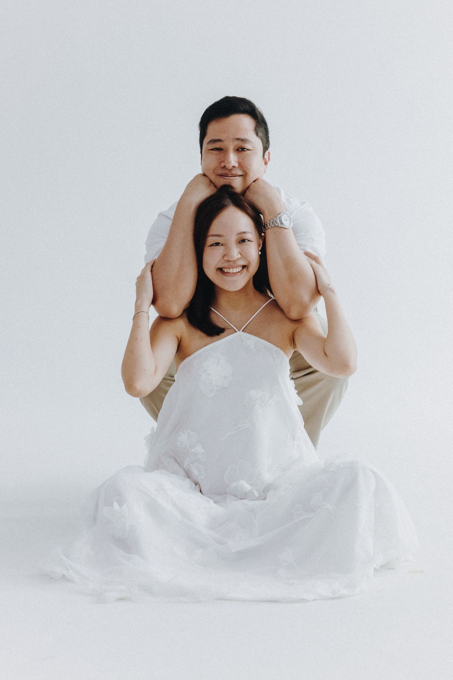 A playful couple poses together in white attire against a simple grey background.