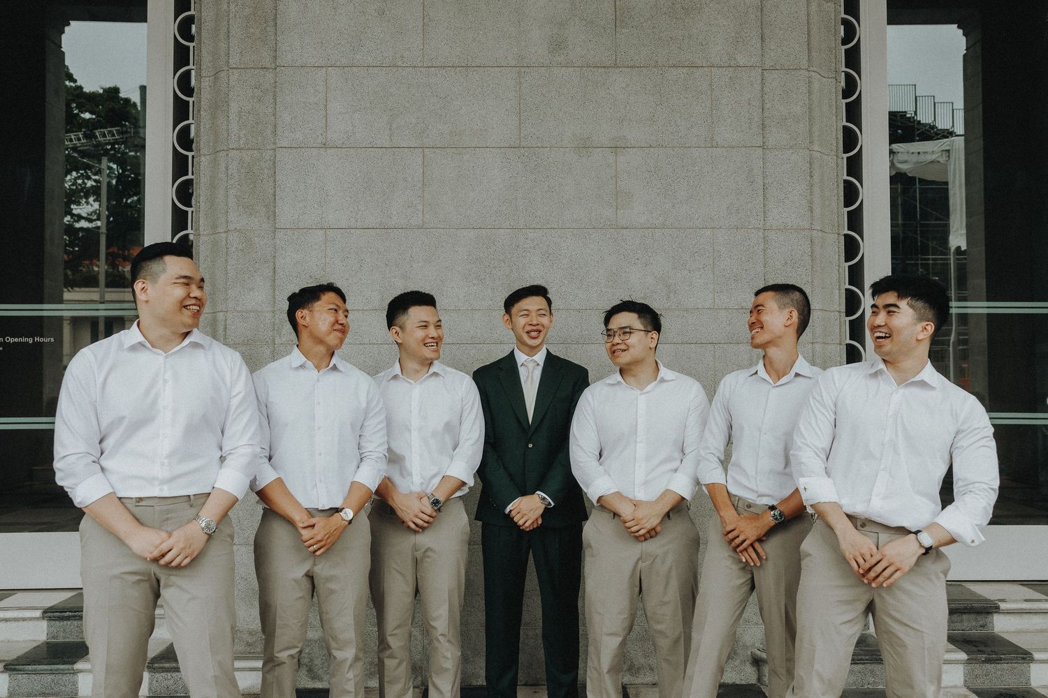 Wedding party members in light colored formal attire lined up against a concrete wall.