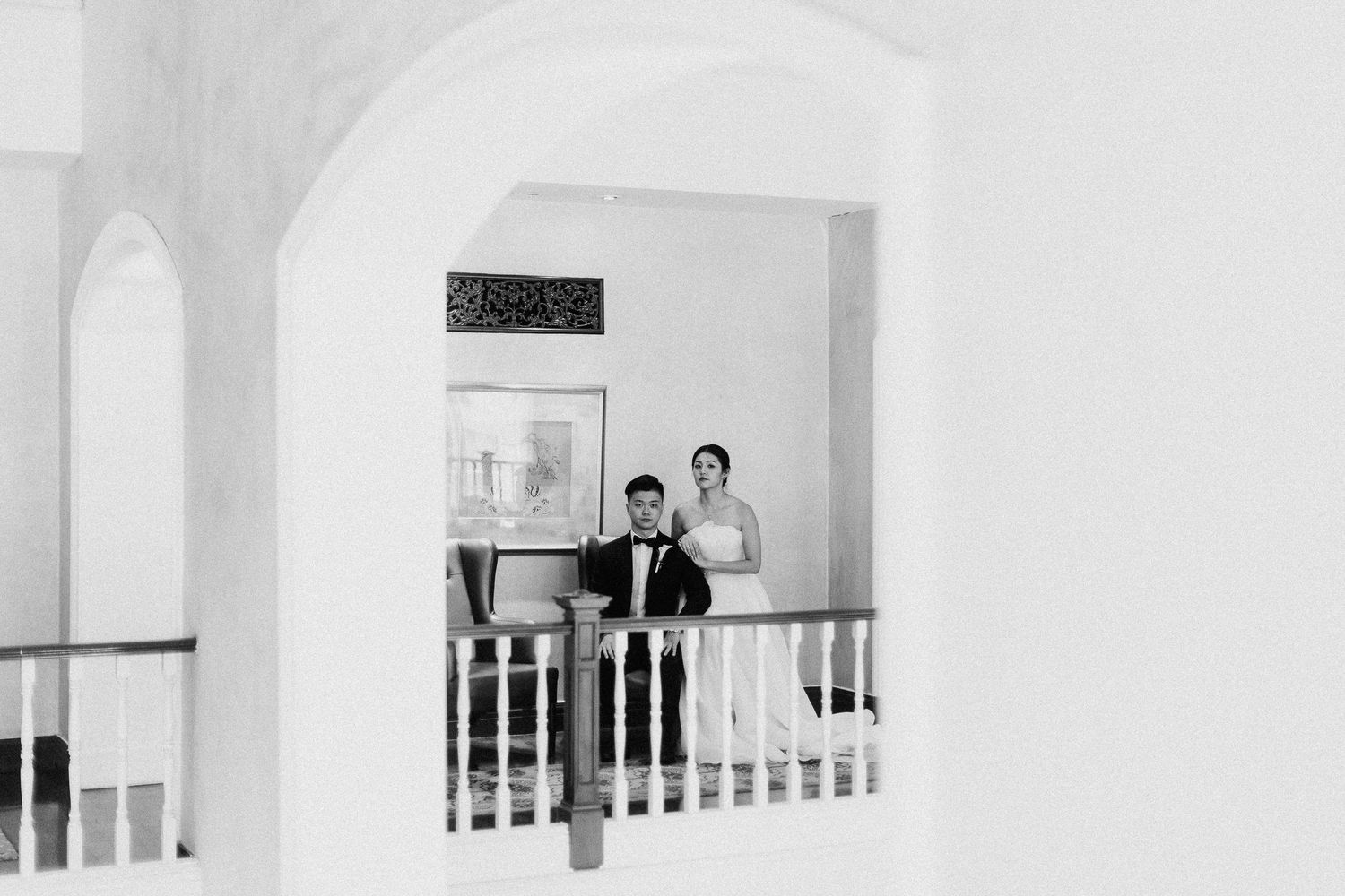 A black and white wedding portrait showing a couple on a balcony with white railings and arched architecture.