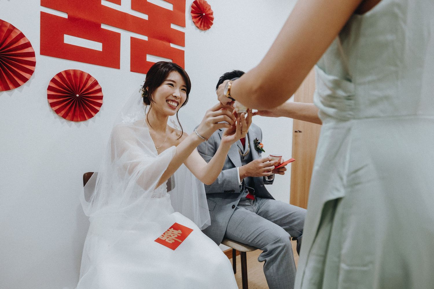 Red paper fan decorations adorn a wall behind a couple during traditional tea ceremony celebrations.