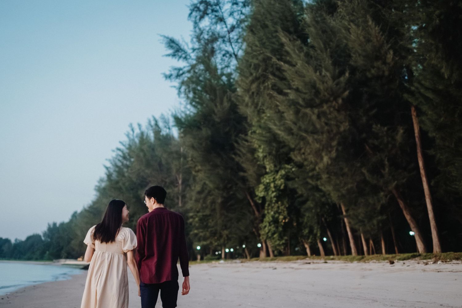A couple walks together along a scenic beach path bordered by tall pine trees at sunset.