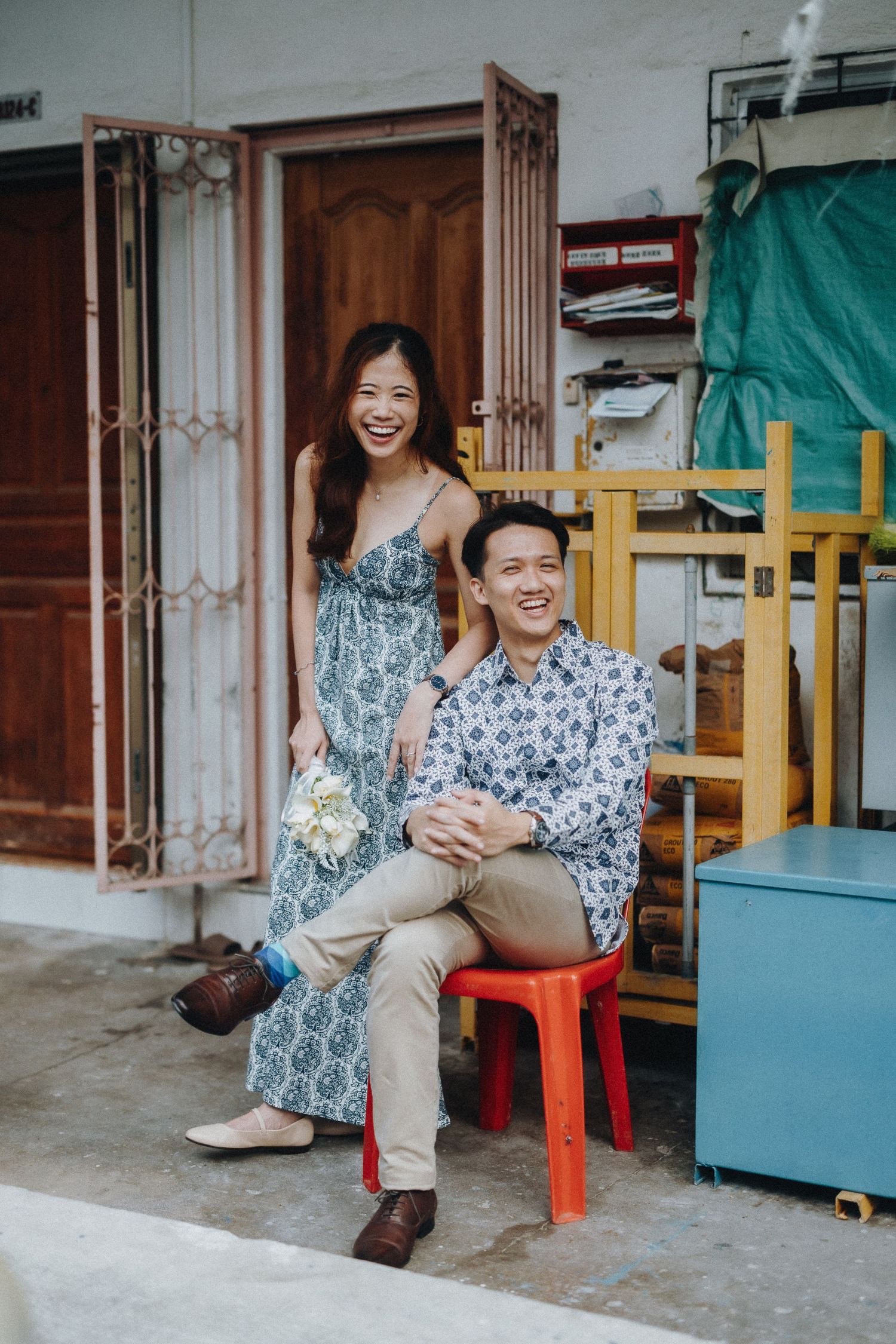 A couple sitting on a red plastic stool shares a candid moment in front of a wooden door and storage shelves.