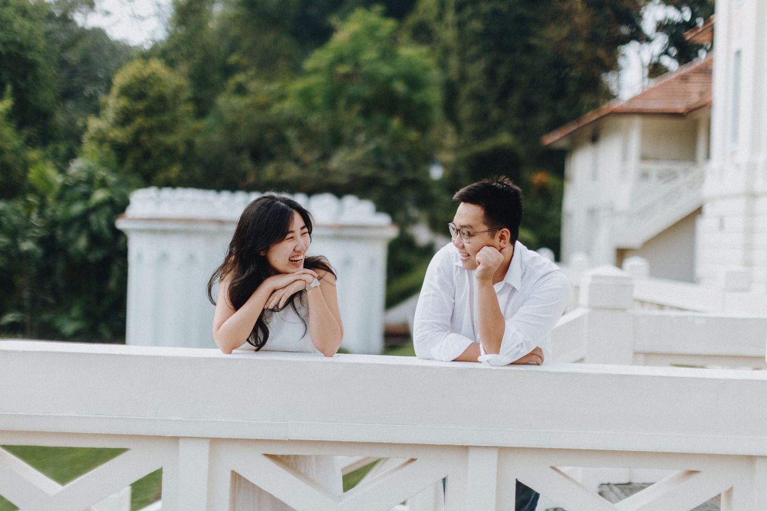 Two people wearing white and black outfits lean on a balcony railing while having an outdoor conversation.