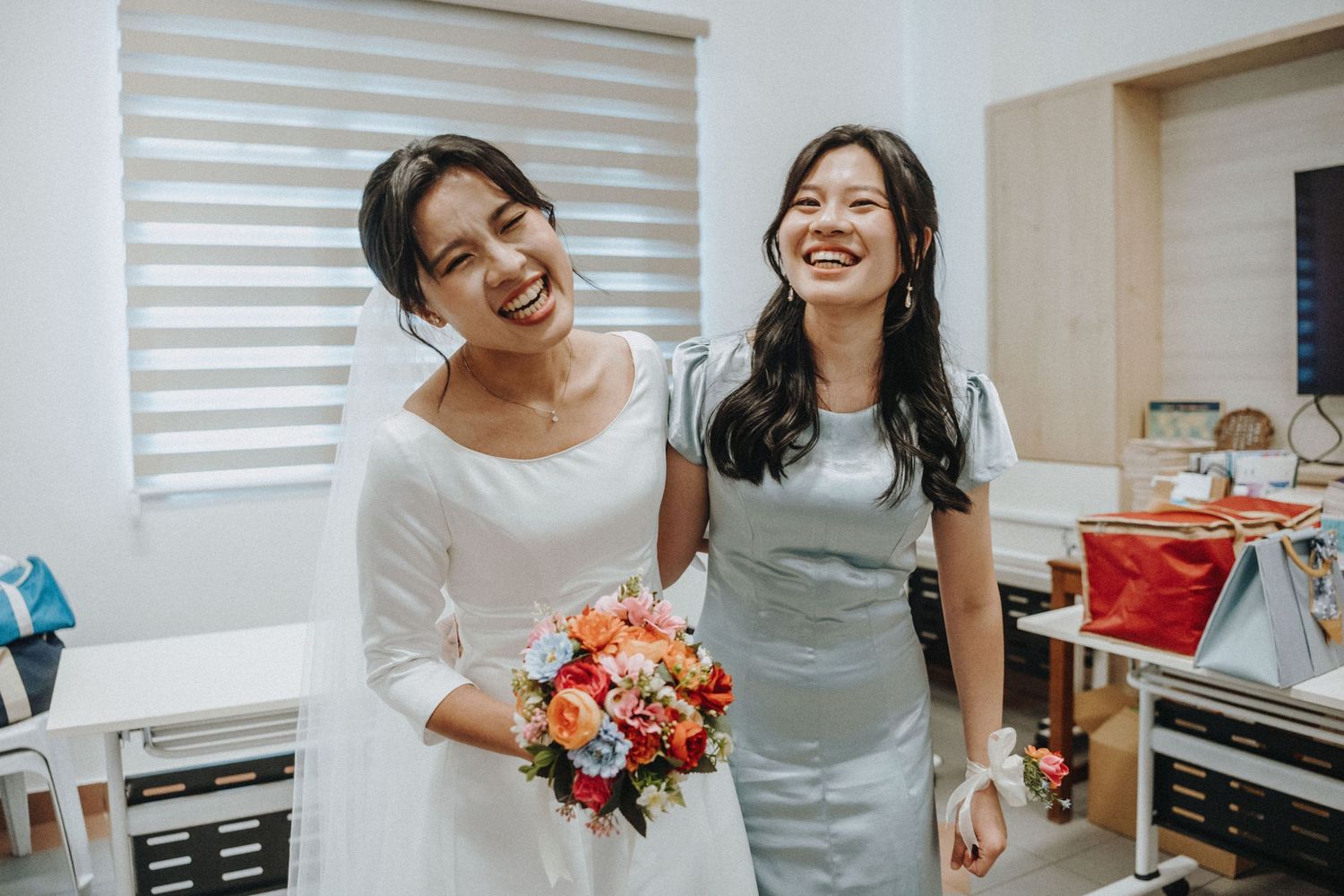 A bride in white dress holding colorful bouquet smiles with bridesmaid in silver dress at wedding.