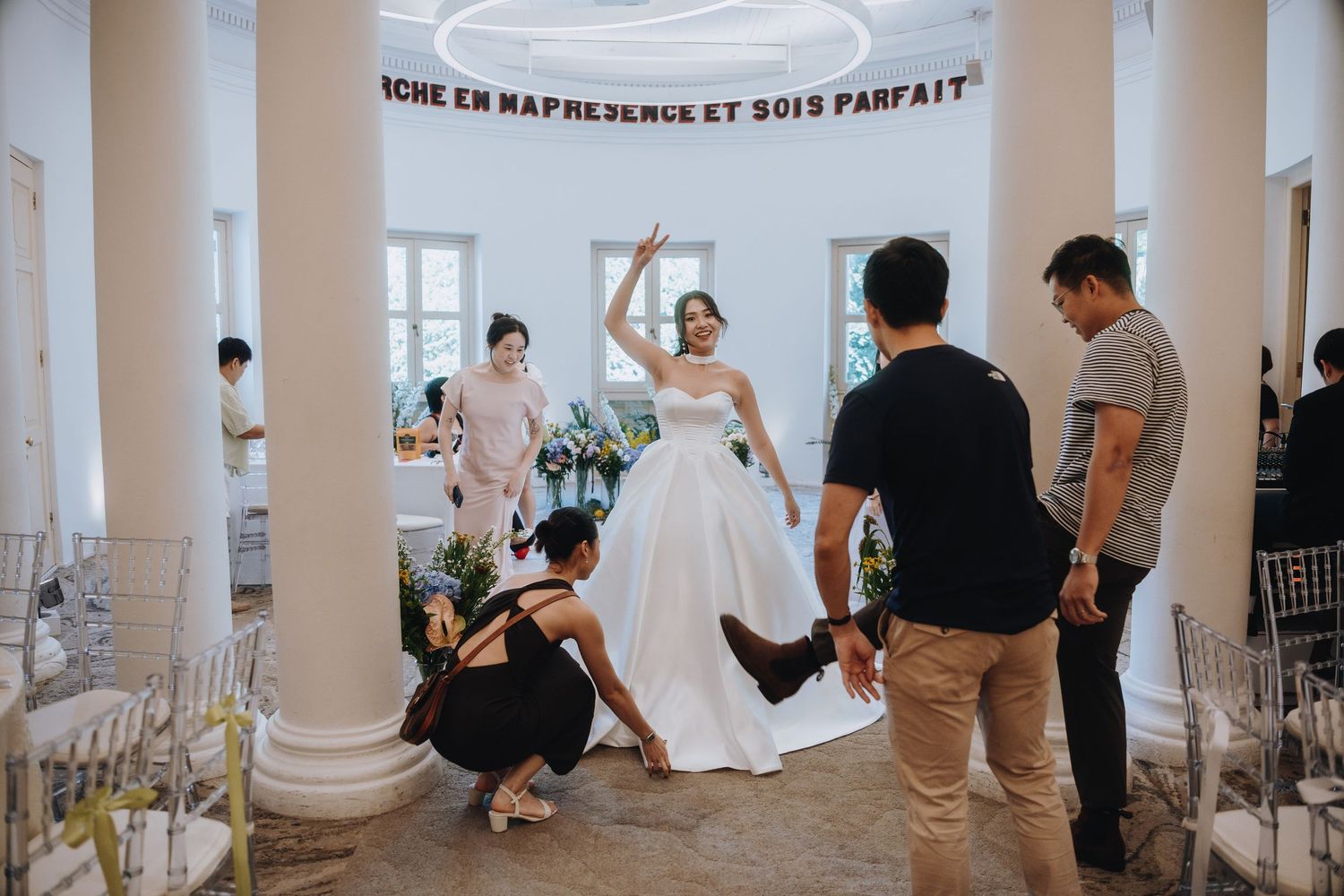 Bride in white gown gets dress adjusted by assistant while posing with guests in elegant venue.