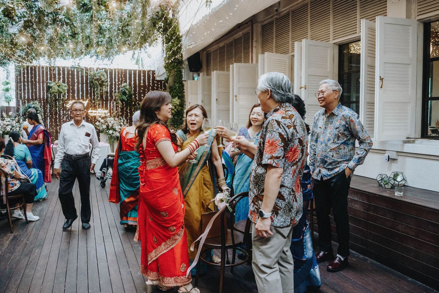 Guests in colorful traditional Indian and Asian attire mingle on a wooden deck at a wedding.