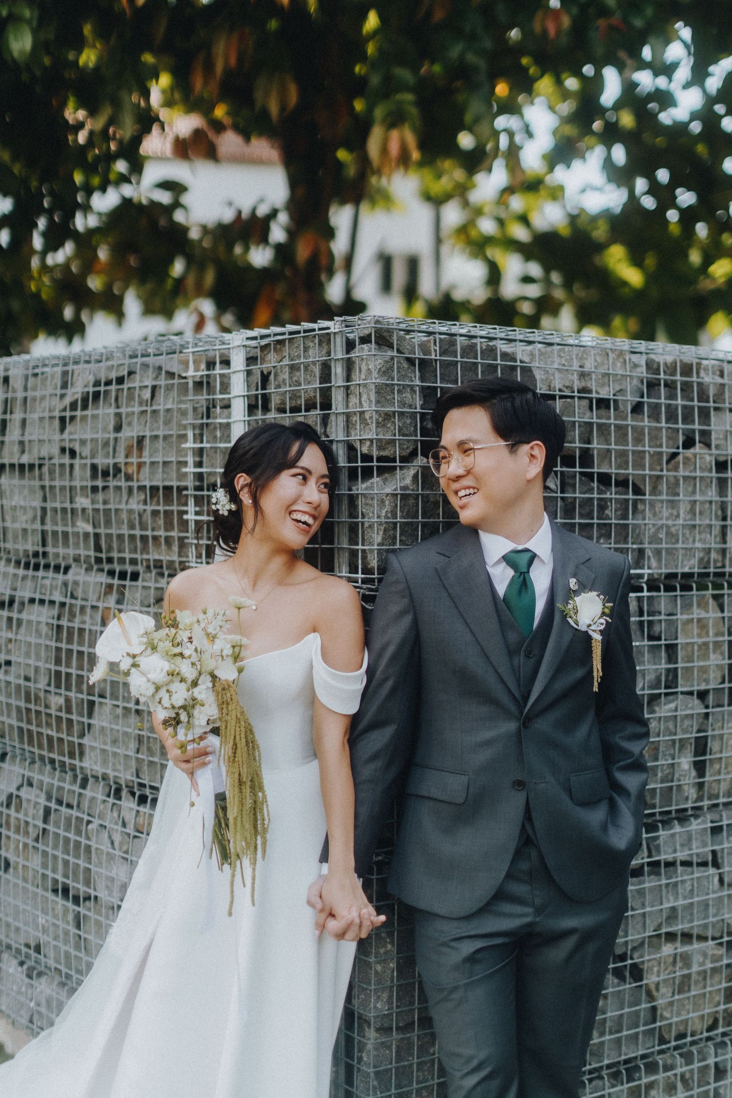 Couple in wedding attire smiling at each other against a stone wall backdrop with lush foliage.