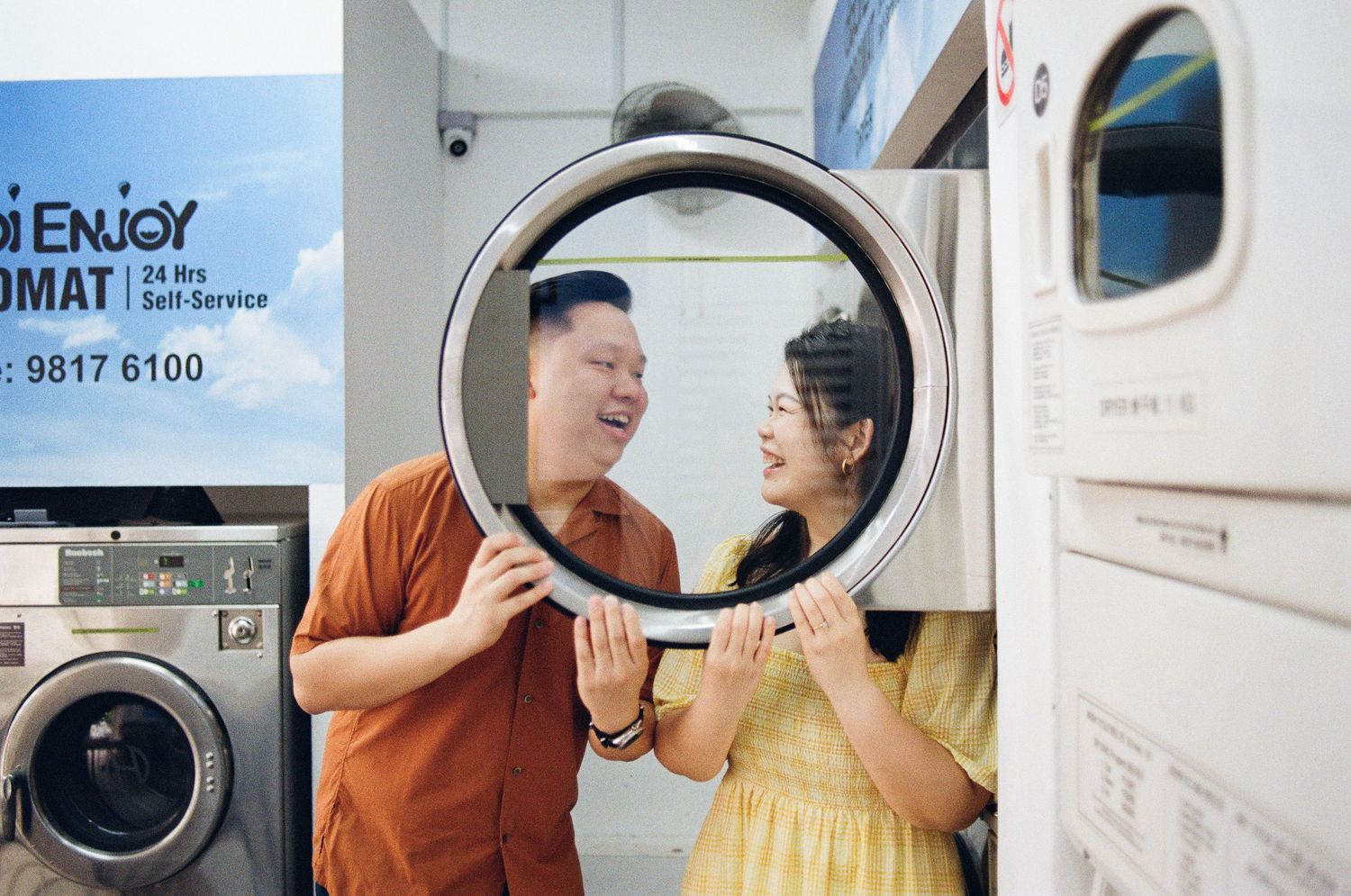 A playful couple poses through a circular washing machine window at a laundromat.