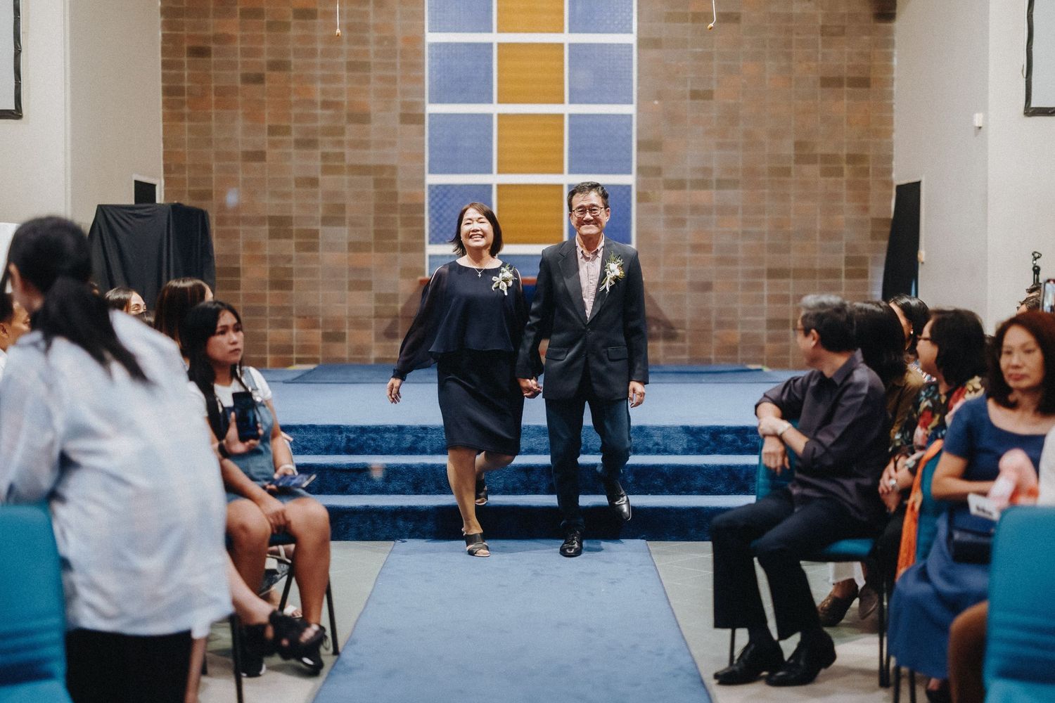 People seated in rows at an indoor venue with brick walls and stained glass windows.