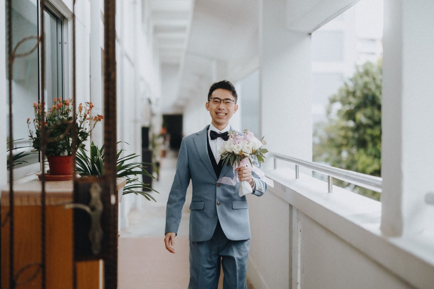 A person in a gray suit holds a floral bouquet while standing in a bright corridor.