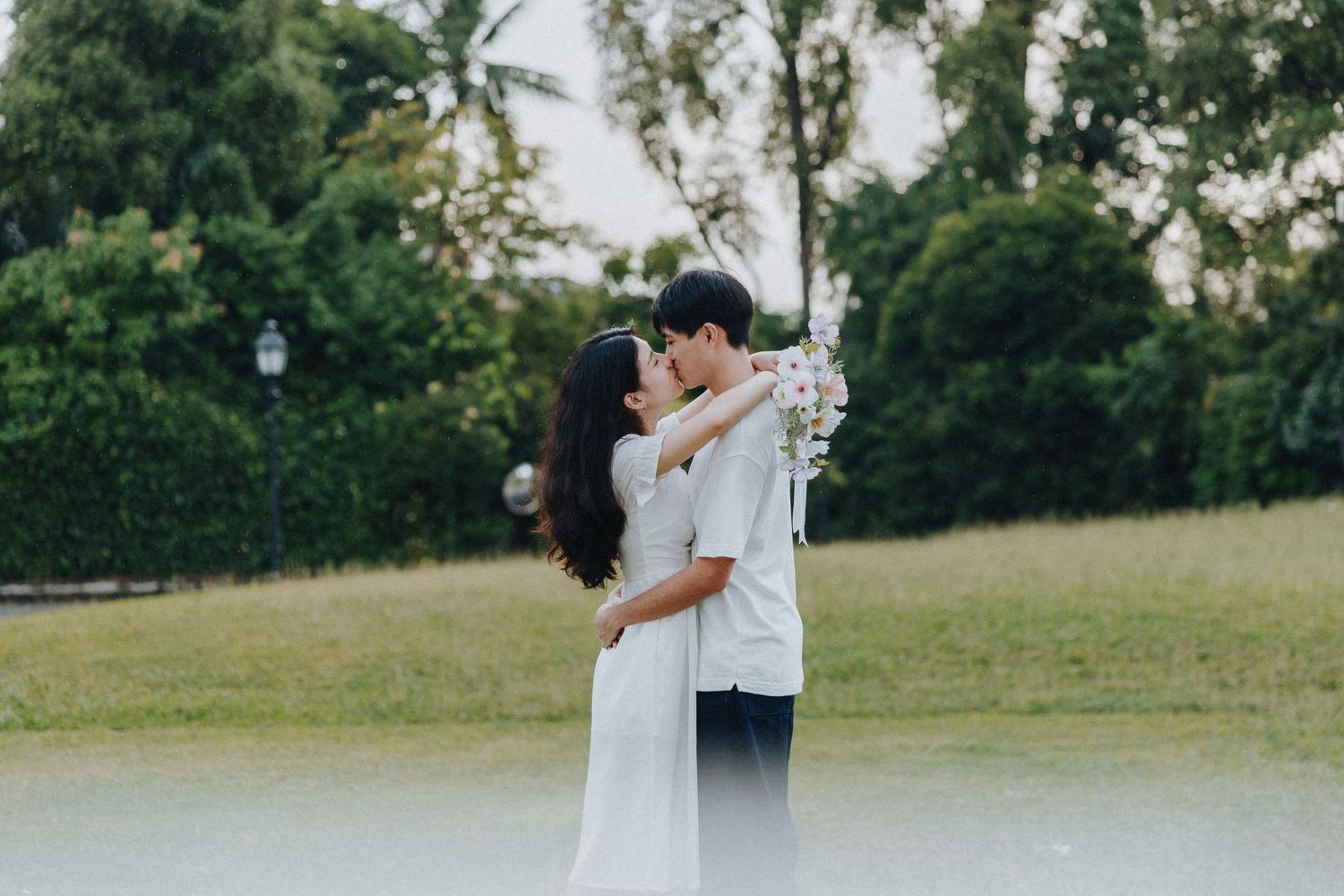 A romantic couple embraces in a white dress and formal attire on a grassy lawn with trees in the background.