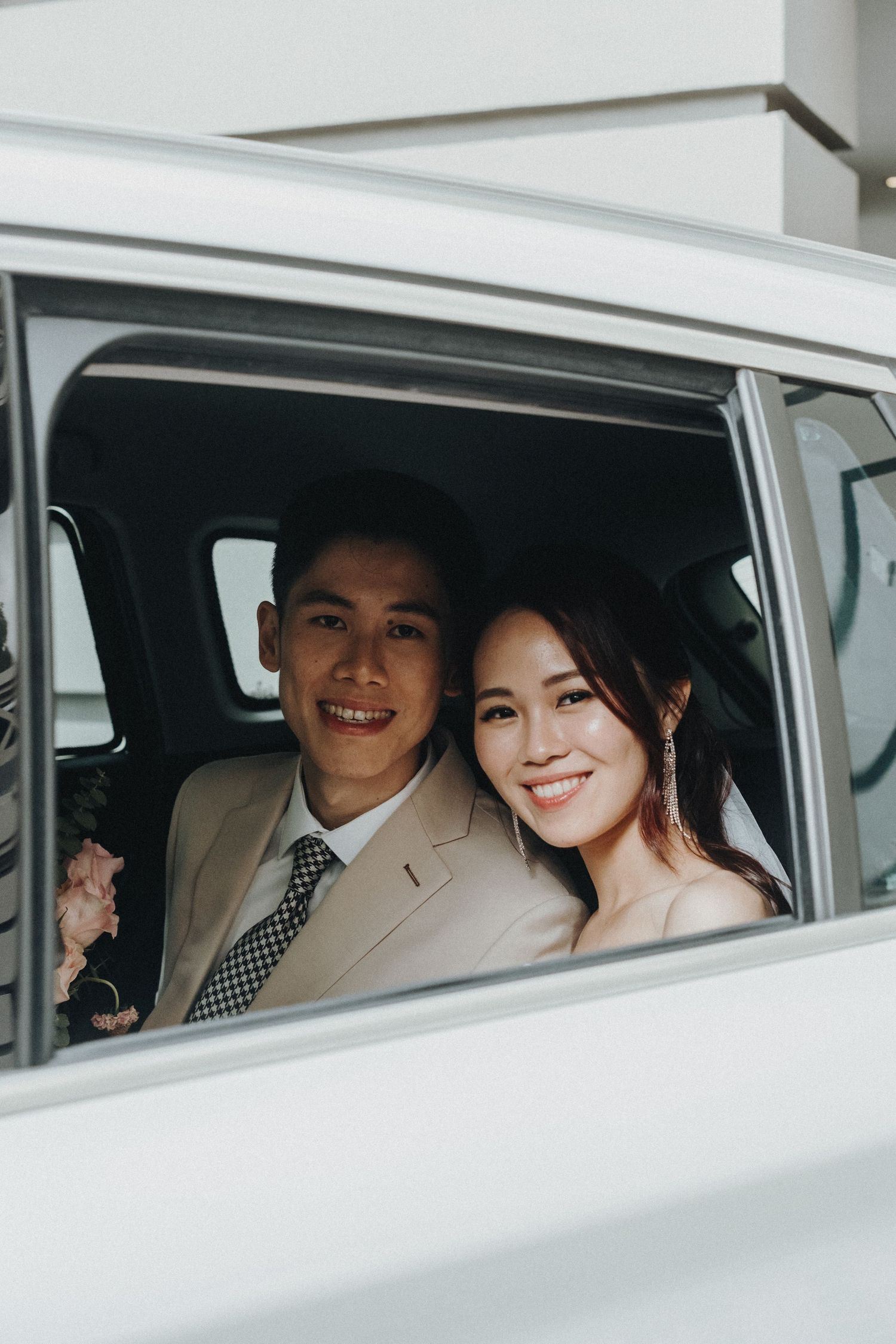 A newly married couple smiles while sitting inside a white classic car during their wedding day.