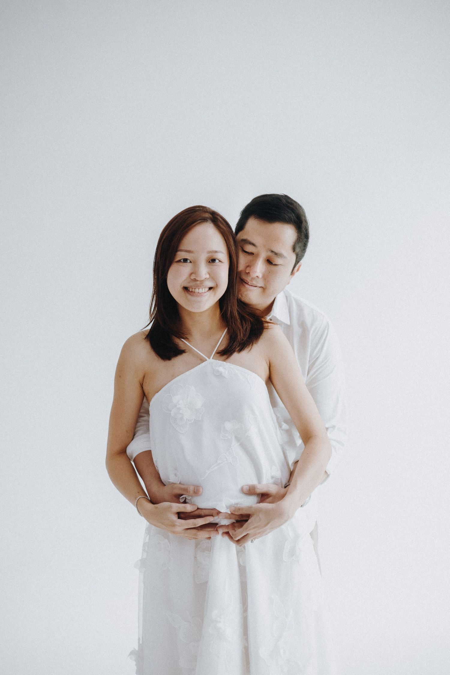 Couple embraces in a romantic maternity photoshoot against white backdrop wearing matching light colored outfits.