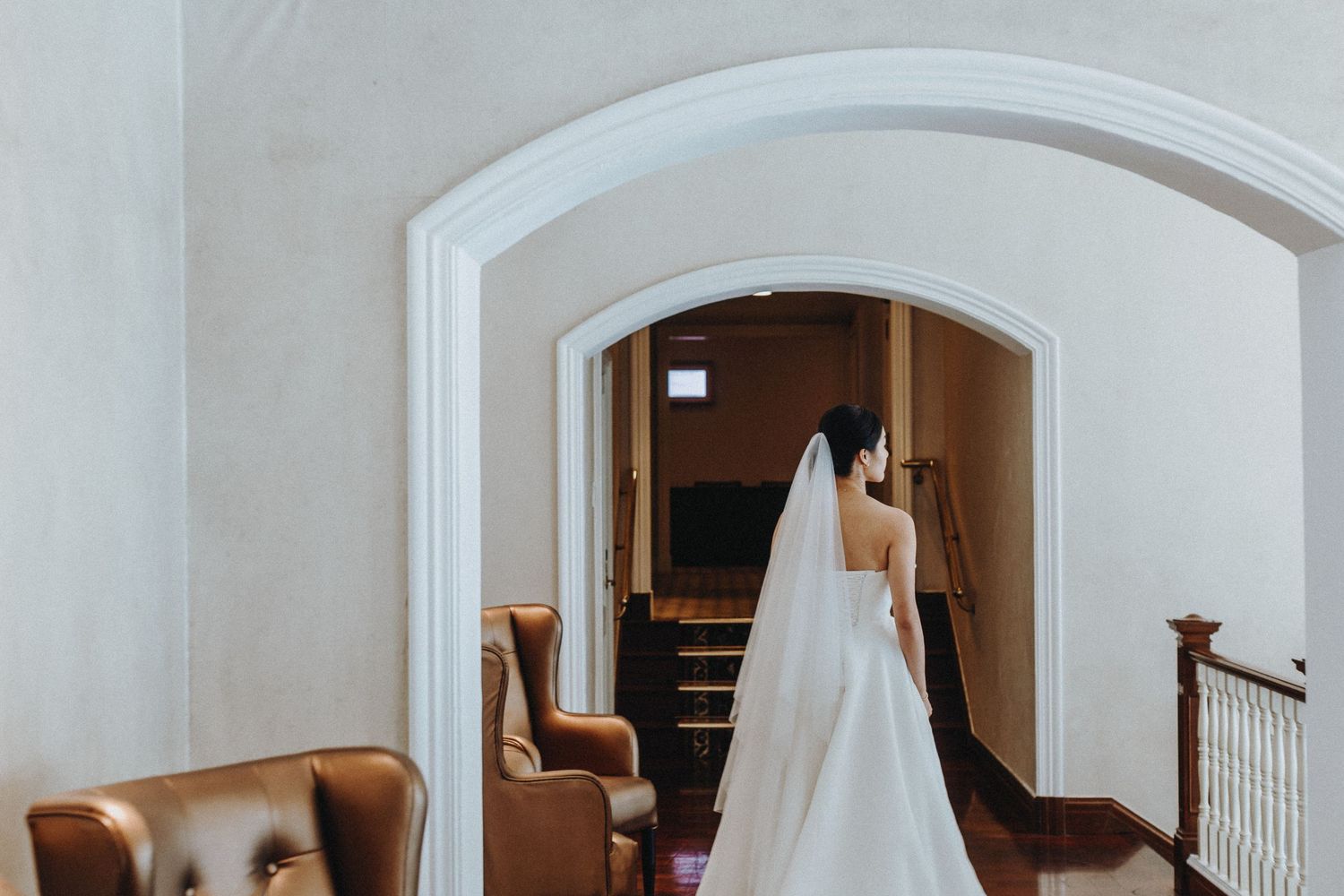 A long white wedding dress seen through an elegant arched doorway with leather chair in the foreground.