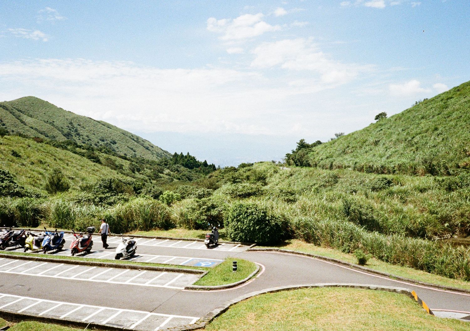 Winding mountain road curves through green hillsides under a bright blue sky with scattered clouds.