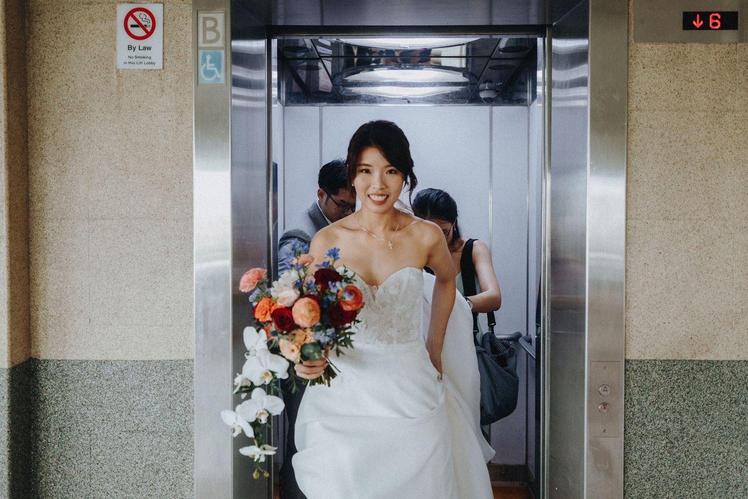 A joyful bride holding a colorful bouquet steps out of an elevator in her white wedding gown.