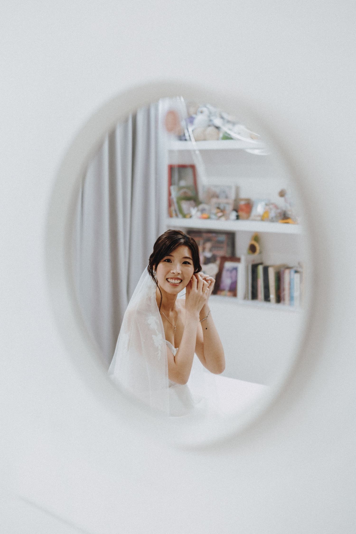 A bridal portrait reflection in a circular mirror showing a woman in a wedding veil with bookshelves in background.