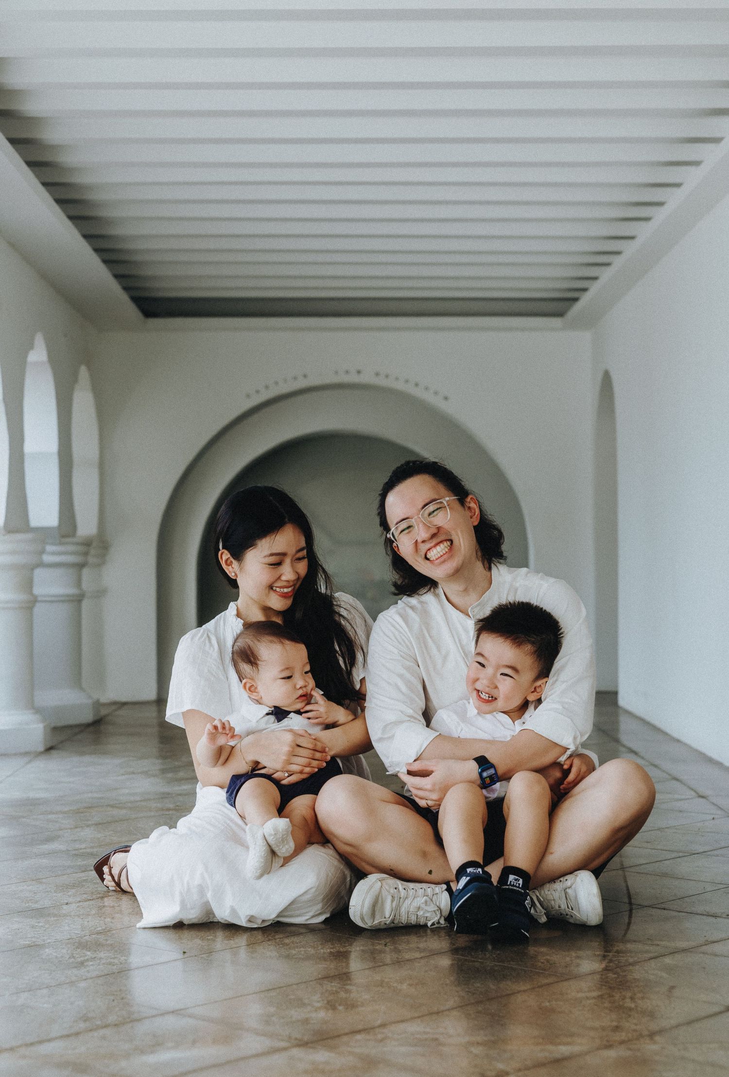A family sits together on a white tiled floor in an elegant hallway with arched columns and natural lighting.